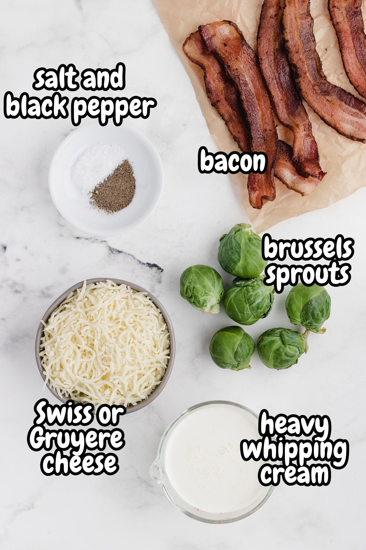 Overhead view of labeled ingredients for Brussels sprouts casserole on a white marble countertop: cooked bacon strips on parchment paper, a small dish of salt and black pepper, six whole Brussels sprouts, a bowl of shredded Swiss or Gruyère cheese, and a glass measuring cup of heavy whipping cream.