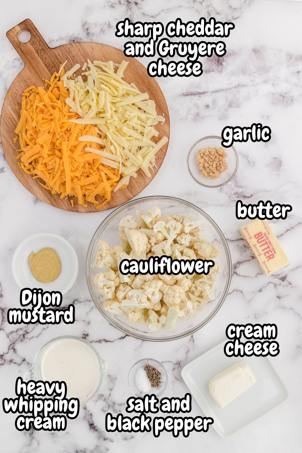 A top-down photo of ingredients for low-carb cauliflower mac and cheese, including cauliflower florets, shredded sharp cheddar and Gruyère cheese, butter, garlic, Dijon mustard, heavy whipping cream, cream cheese, salt, and black pepper arranged on a white marble surface.