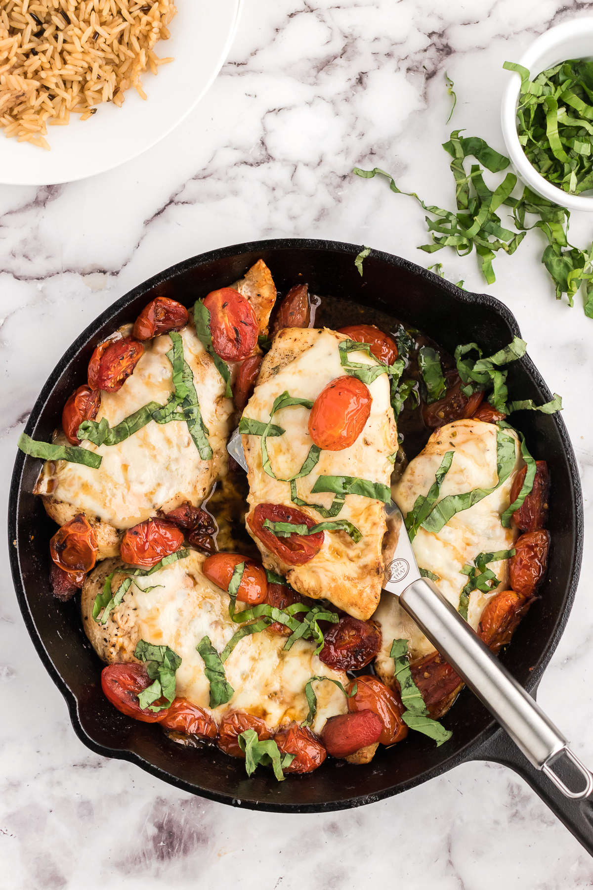 the baked caprese chicken in a skillet with one chicken breast topped with balsamic and basil on a spatula.