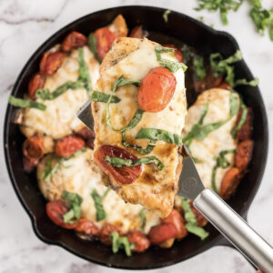 the baked caprese chicken in a skillet with one chicken breast topped with balsamic and basil on a spatula.