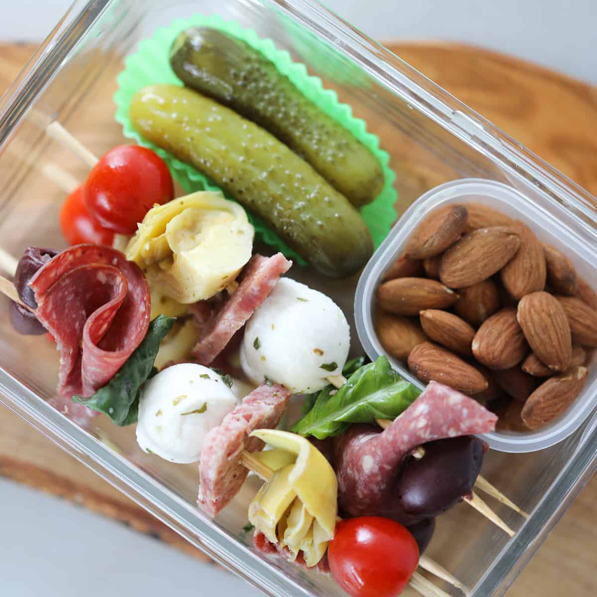 the antipasto skewers keto recipe makes a great low carb lunch, shown in a meal prep conntainer with nuts and pickles