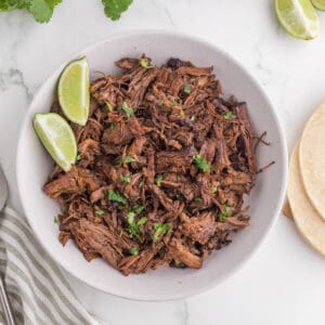 A bowl of fresh made barbacoa beef with a side of lime wedges.
