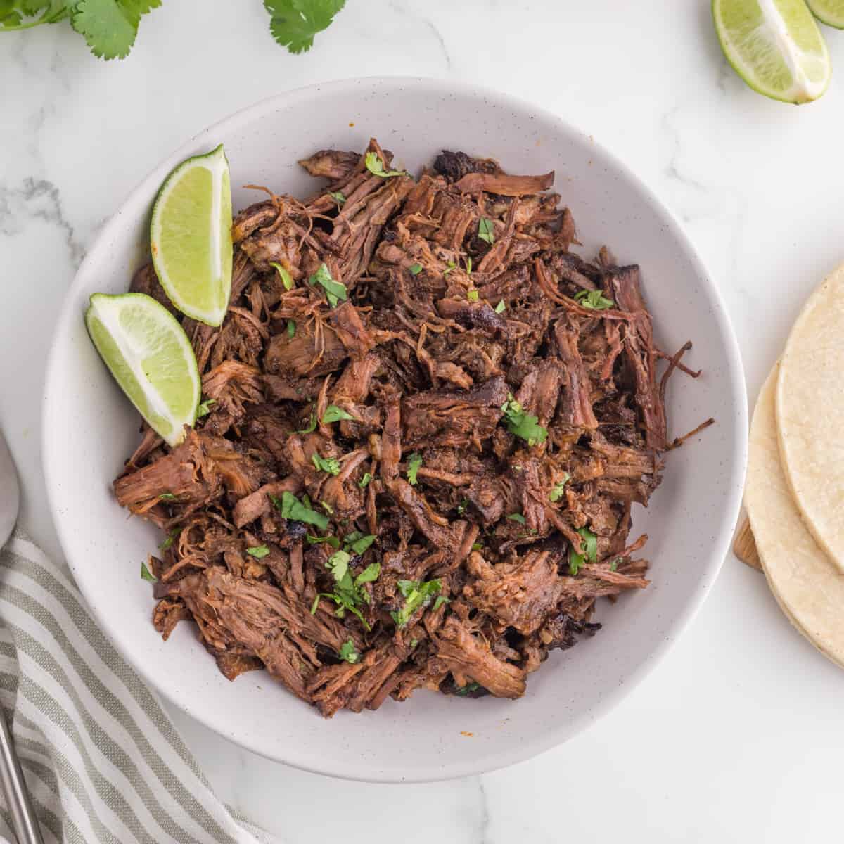 A bowl of fresh made barbacoa beef with a side of lime wedges.