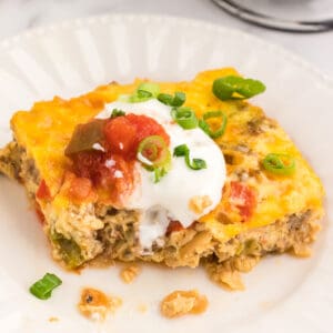 A square slice of keto breakfast casserole on a white plate topped with sour cream and salsa.