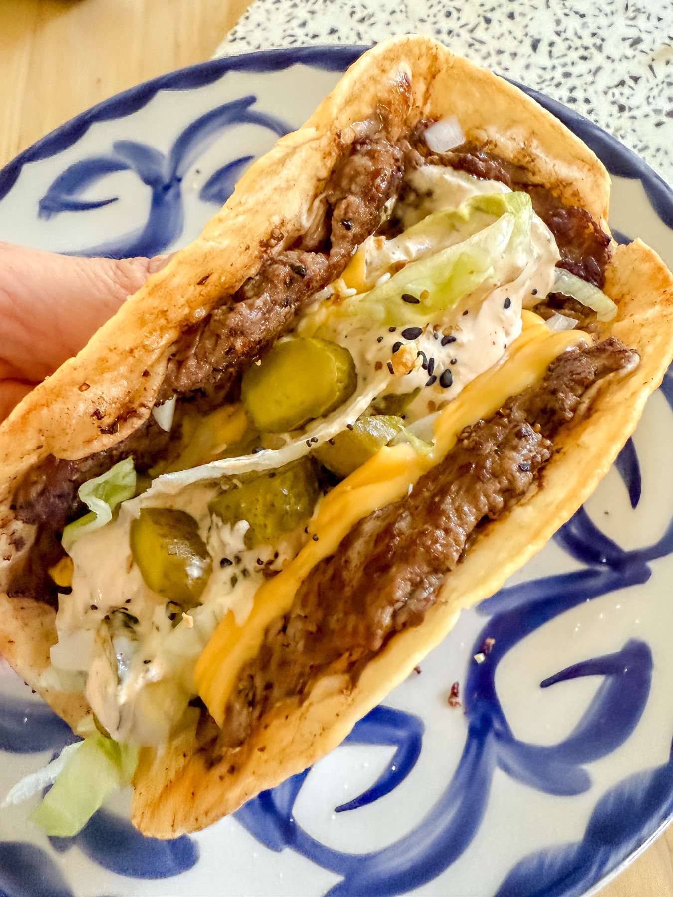 blackstone big mac smash tacos in a persons hand folded over ready to eat on a blue and white plate